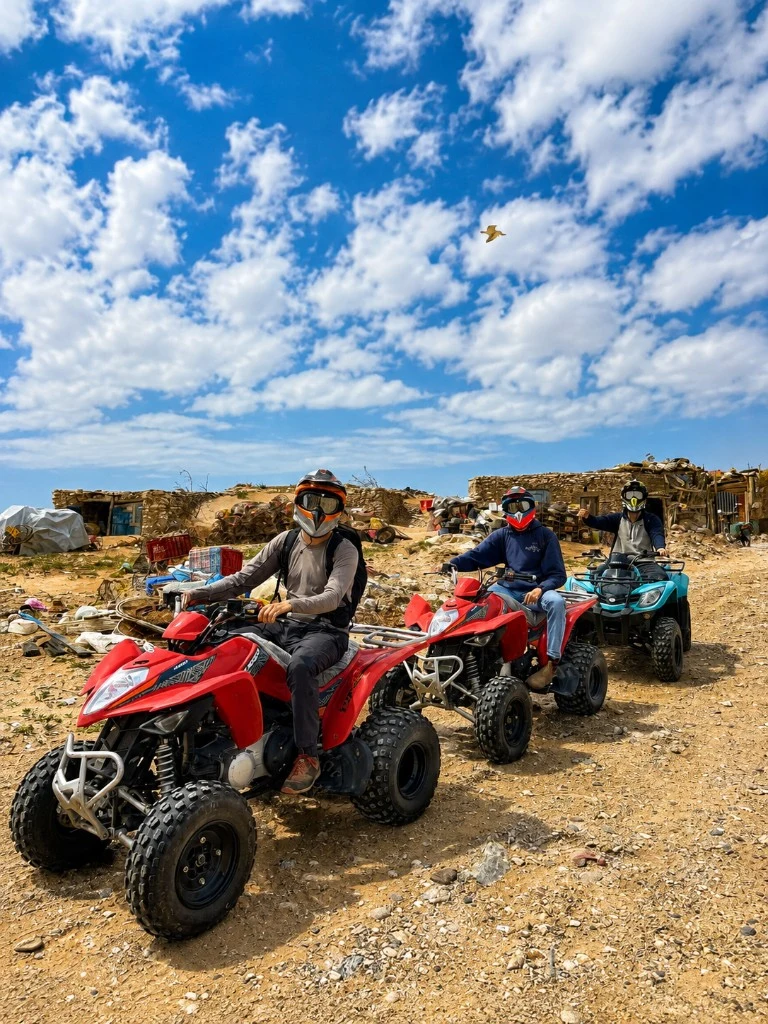 Three riders on red and turquoise quad bikes on a rocky Essaouira track beside a stone rural house